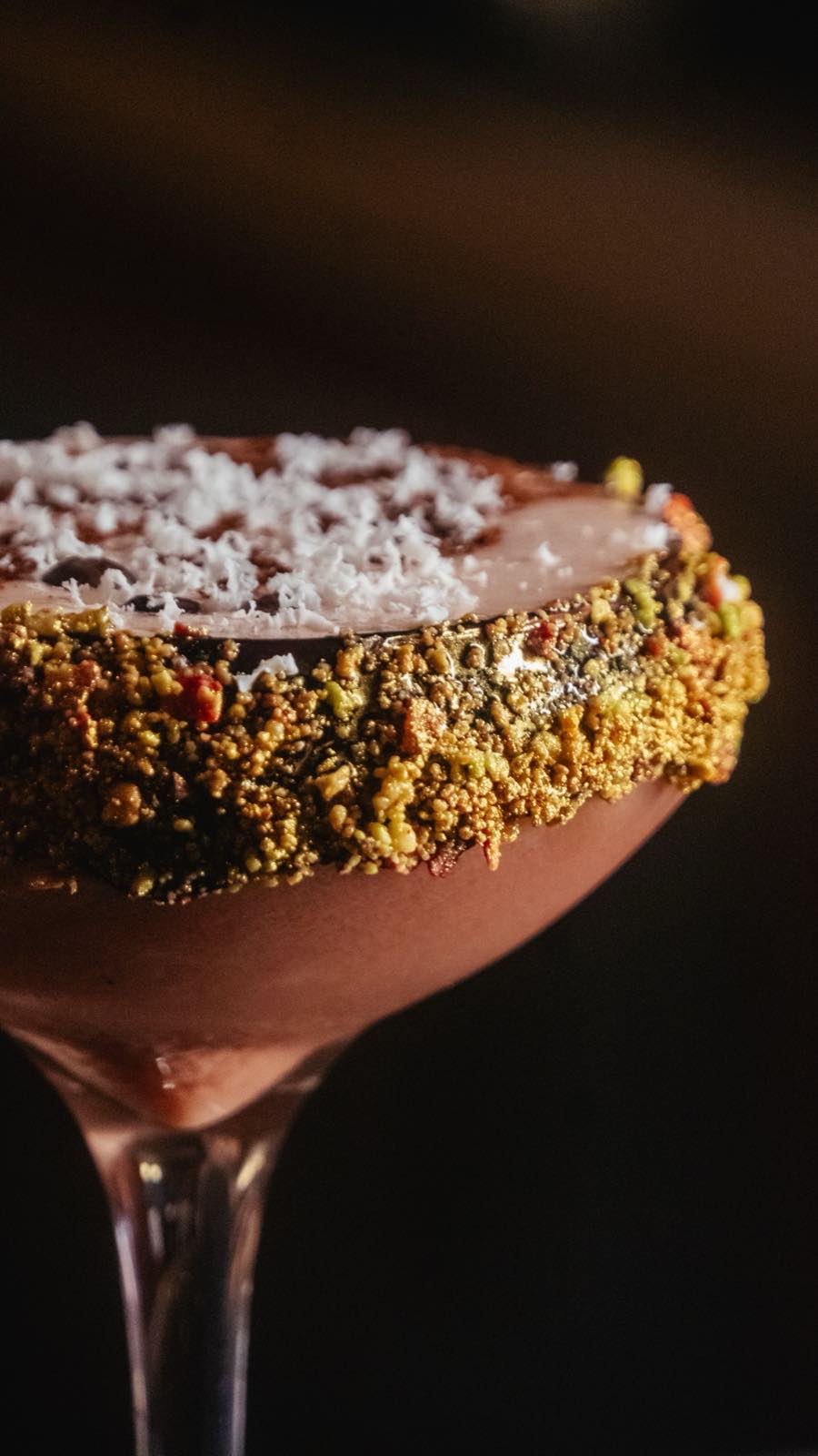 A cocktail in a coupe glass with a salted, herb-crusted rim, lit warmly against a dark background