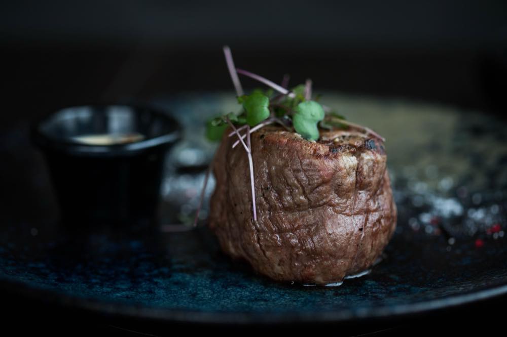 A filet of beef on a dark plate with a small jug of sauce